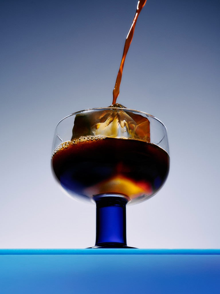 A splash of rich, dark coffee pours into a clear glass on a blue surface
