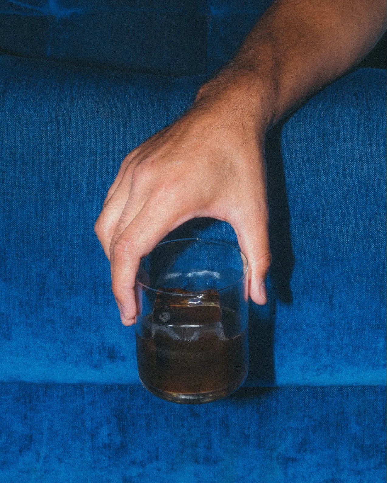 A hand holds a glass filled with a dark beverage and ice cubes against a blue sofa